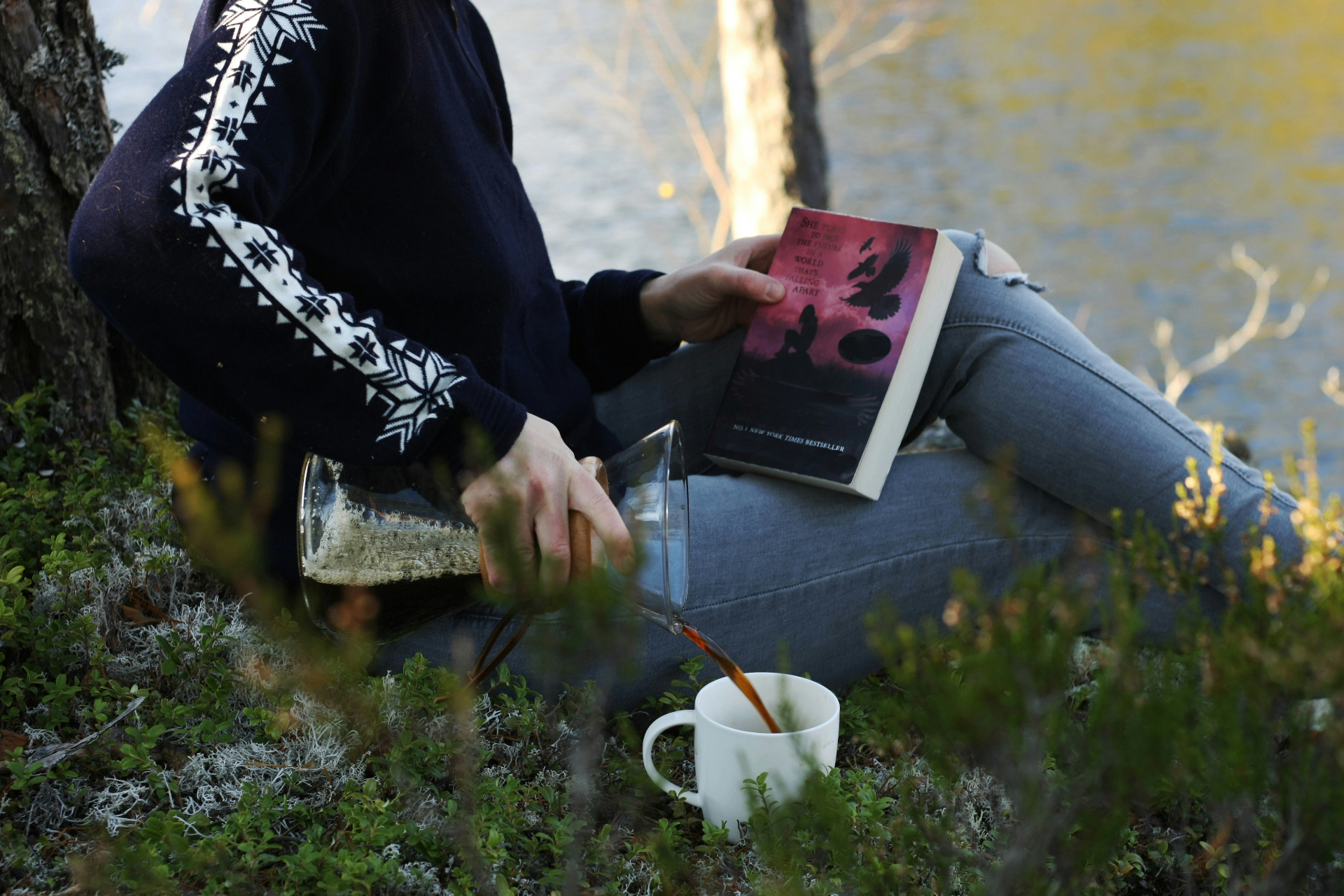 Woman by a lakeside pouring coffee while holding a book — quiet morning ritual